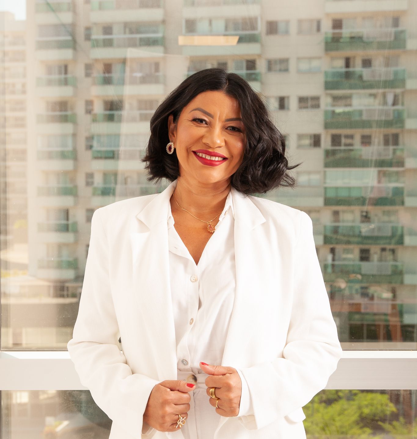 Dra. Neuma Lopes, biomédica, de terno branco, sorrindo diante da janela